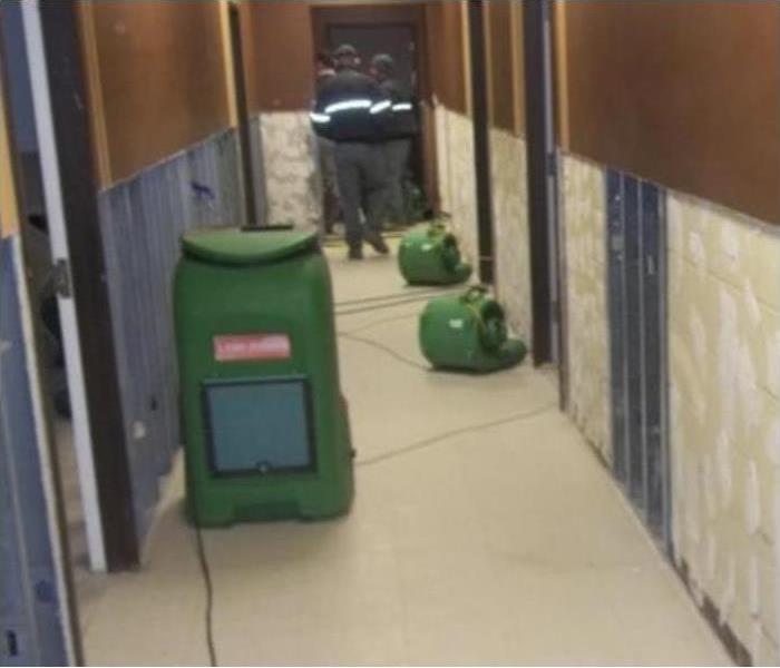 Air movers and dehumidifiers placed in a hallway and at the end of the hallway there are three technicians